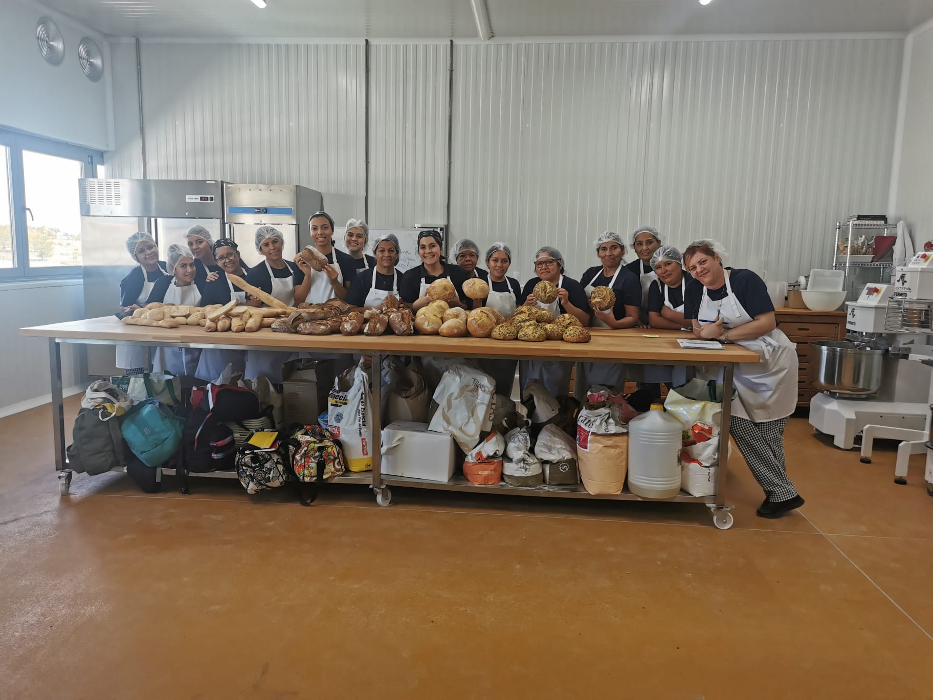 Un grupo de mujeres con delantal y gorro protector posando detrás de una mesa de obrador con diferentes panes