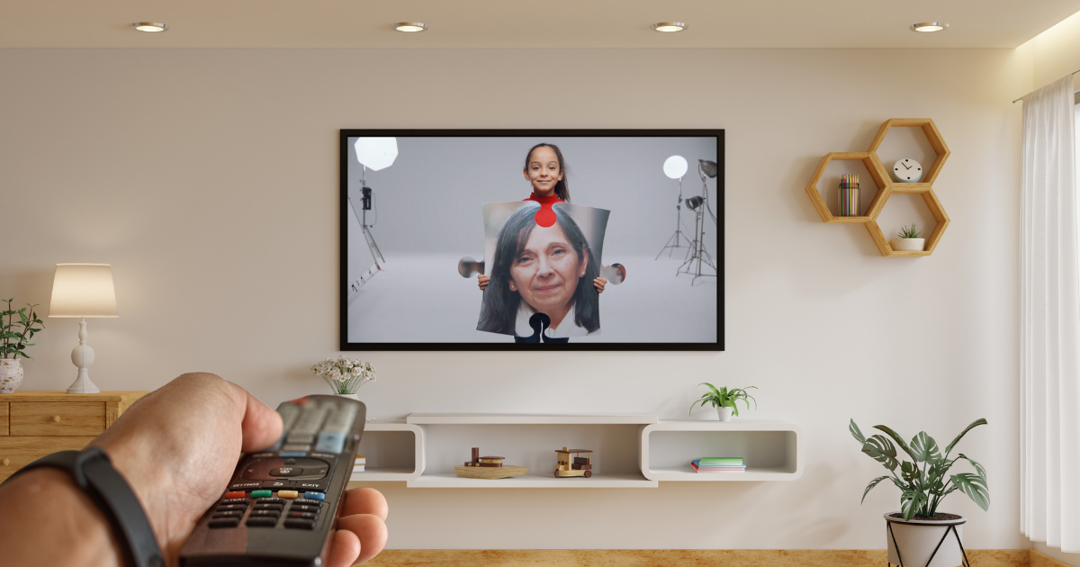 Un salón de una casa con un televisor donde se ve una de las imágenes de la campaña ContigoTodoEncaja. En primer plano, una mano sujetando un mando de tv.