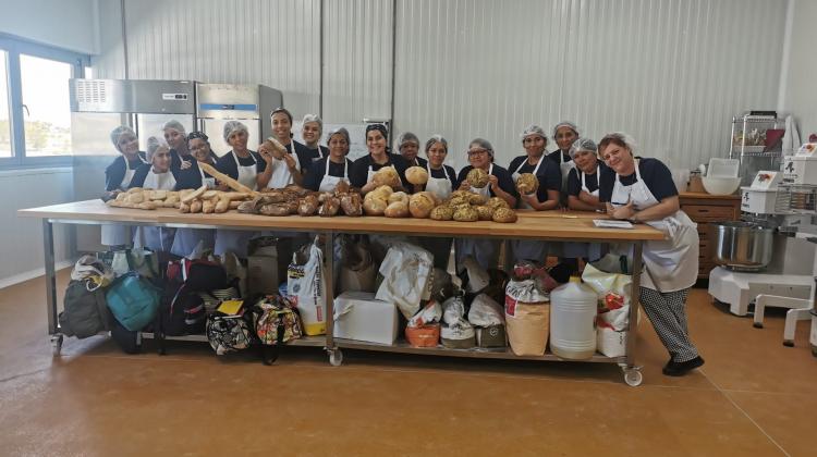 Un grupo de mujeres con delantal y gorro protector posando detrás de una mesa de obrador con diferentes panes